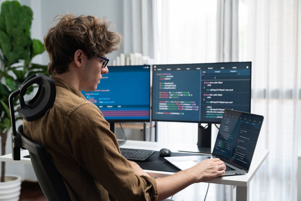 Softwareentwickler schreibt Programmcode an einem Arbeitsplatz mit drei Bildschirmen